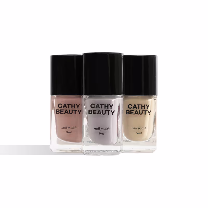 Três frascos de esmalte da marca Cathy Beauty, com embalagens pretas e rótulos com o nome da marca. As cores dos esmaltes são um tom rosado, branco e bege claro.