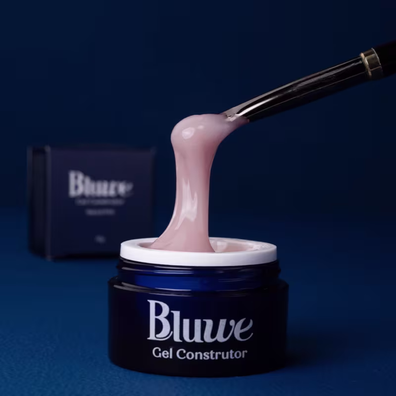 A imagem mostra o gel construtor Bluwe, em fundo azul escuro. É um frasco azul escuro com gel rosa. Há o texto "Bluwe Gel Construtor" em letras brancas e um aplicador de gel metálico longo e fino mostrando a consistência e textura do produto.