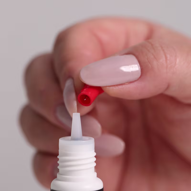 O aplicador possui um tubo branco com uma ponta vermelha, provavelmente para facilitar a aplicação do produto nos cílios. O design é simples e prático, com foco na funcionalidade do aplicador.