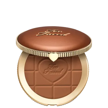 Pó bronzeador compacto com acabamento aveludado em embalagem redonda de metal dourado com detalhes em relevo e coração no centro. Apresenta textura e tonalidade marrom escura com inscrição "Too Faced" na superfície.