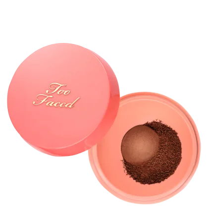 Pó solto Too Faced aberto sobre fundo branco. À esquerda, a tampa redonda e rosada exibe "Too Faced" em letras douradas. À direita, a base rosada mostra o pó solto em tom marrom-escuro ao redor do dosador "Peach Pit™"
