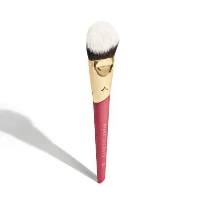 A imagem mostra um pincel de maquiagem, visto de um ângulo um pouco mais elevado, sem ser exatamente de frente. Com cabo vermelho e dourado, e cerdas pretas com ponta branca. Na parte vermelha, logo da marca e linha do produto escrito em dourado.