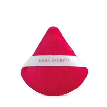 A imagem mostra uma esponja de maquiagem triangular de cor vermelha com uma fita branca na parte central, com  o nome da linha, Niina Secrets.