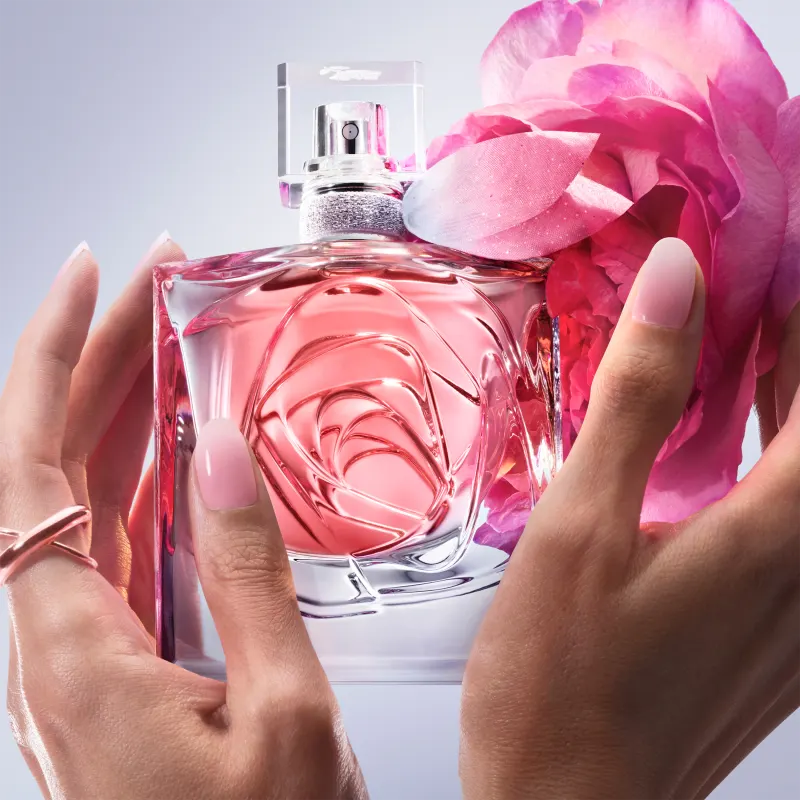 Frasco de perfume Lancôme com design floral em vidro transparente, tampa prateada e detalhe em rosa. Embalagem elegante e sofisticada em tons de rosa e branco.