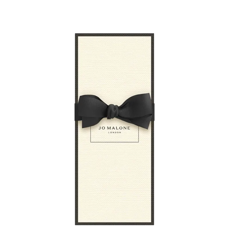 Caixa retangular de papelão branco com bordas pretas, contendo o logotipo "Jo Malone London" impresso. Um laço preto de cetim adorna a parte frontal da caixa.