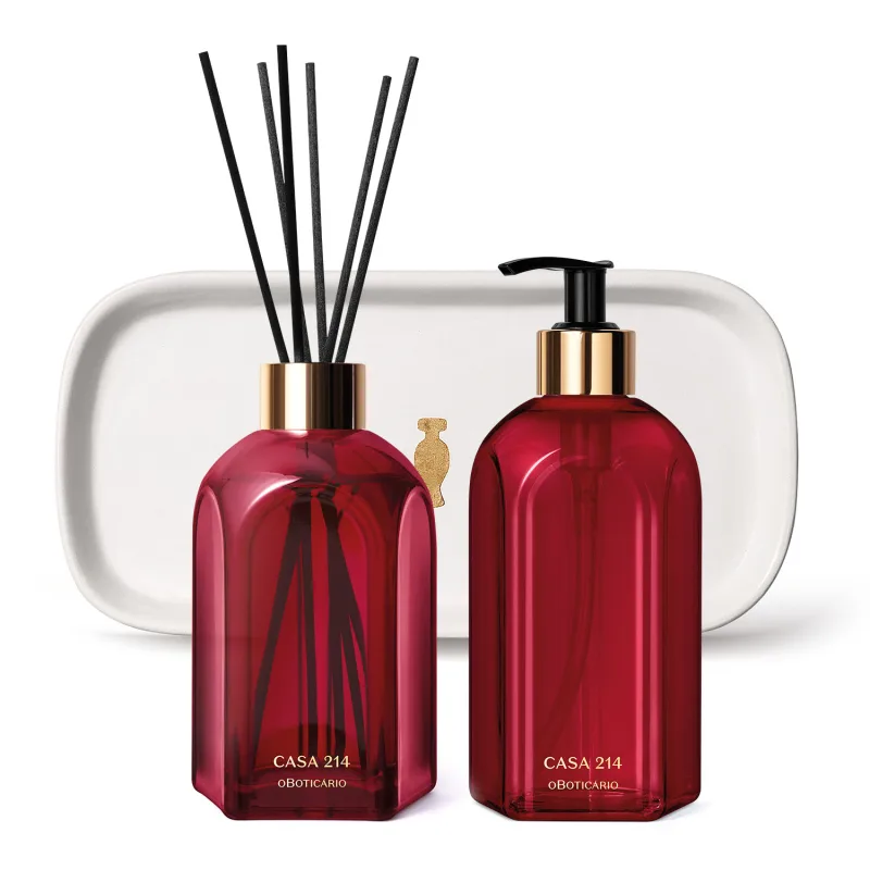 Três produtos da marca Casa 214 Gardênia Imperial: dois vidros vermelhos, sendo um difusor de ambientes com valetas e o outro com bico dosador preto de sabonete líquido e uma badeja de lavabo branca com logo da marca O Boticário em dourado.