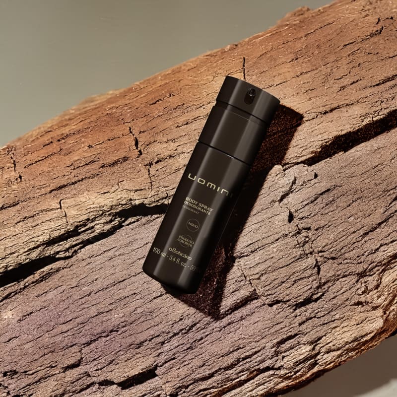 Imagem de embalagem preta de body spray da marca Uomini sobre superfície de madeira rachada, em cenário com tons terrosos e sombras.