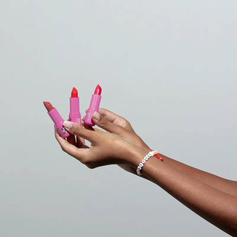 Três batons com embalagem e cores diferenes sendo segurados por mãos femininas negras contra fundo branco. No braço da modelo, vemos uma pulseira de miçangas brancas com detalhes coloridos.