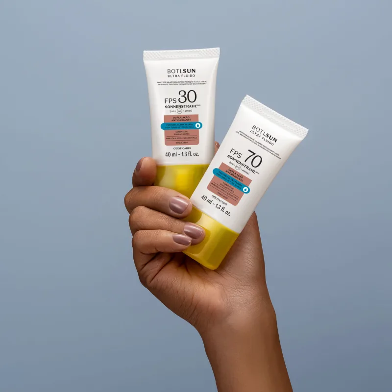 Mão segurando dois frascos brancos com tampas amarelas, letras pretas e detalhes nas cores azul e laranja contendo protetor solar facial antioxidante FPS 70 Ultra Fluído da marca Boti.Sun em fundo azul celeste.