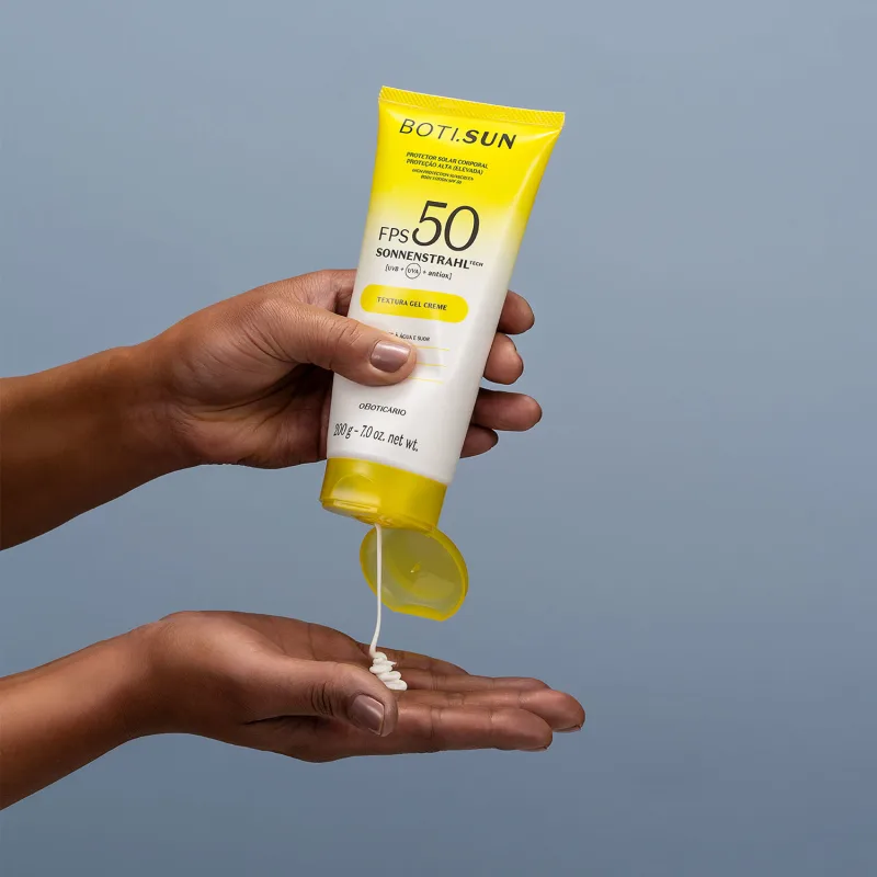 Mão segurando uma Bisnaga amarela e branca com tampa amarela e letras pretas contendo protetor solar corporal FPS 50 da marca Boti.Sun e jogando um pouco do creme branco na outra mão em fundo azul celeste.