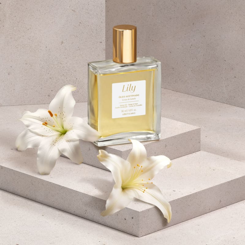 Um vidro quadrado com líquido bege, tampa dourada, rótulo branco com letras bege de óleo acetinado multifuncional corpo e cabelo da marca Lily está em uma plataforma branca, com folhas brancas ao redor, em um cenário com fundo cinza.