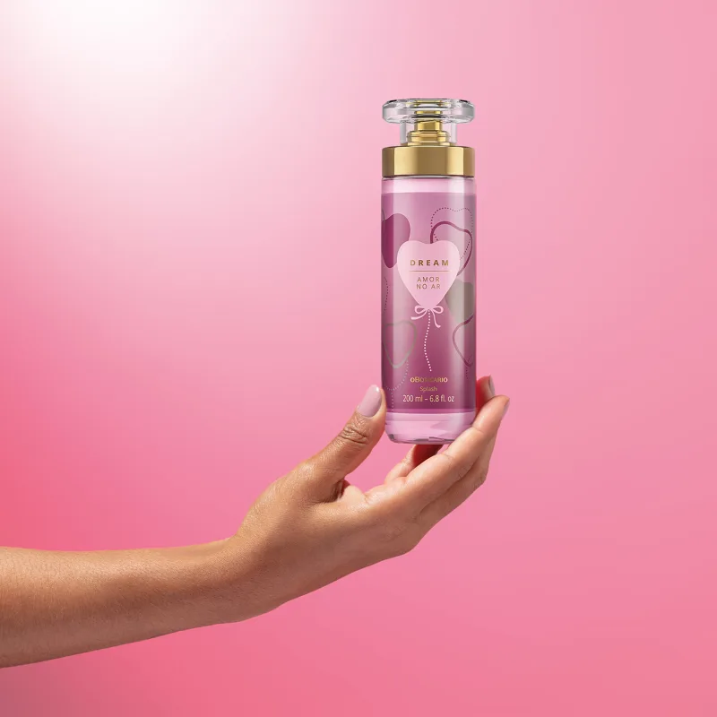 Imagem de uma mão segurando um frasco de body splash Dream Amor no Ar. A embalagem conta com tampa transparente, válvula spray dourada e rotulo em tons de roxo com coração ilustrativo . O cenário traz um fundo em tons de lilás e branco.