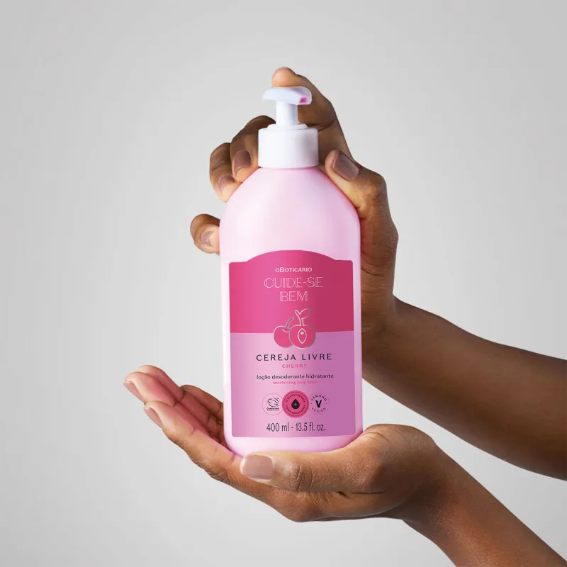 Mão feminina segurando um frasco branco com bomba dosadora e rótulo nas cores rosa e lilás com cerejas ilustradas contendo loção hidratante corporal Cuide-se Bem Cereja Livre em fundo lilás claro.