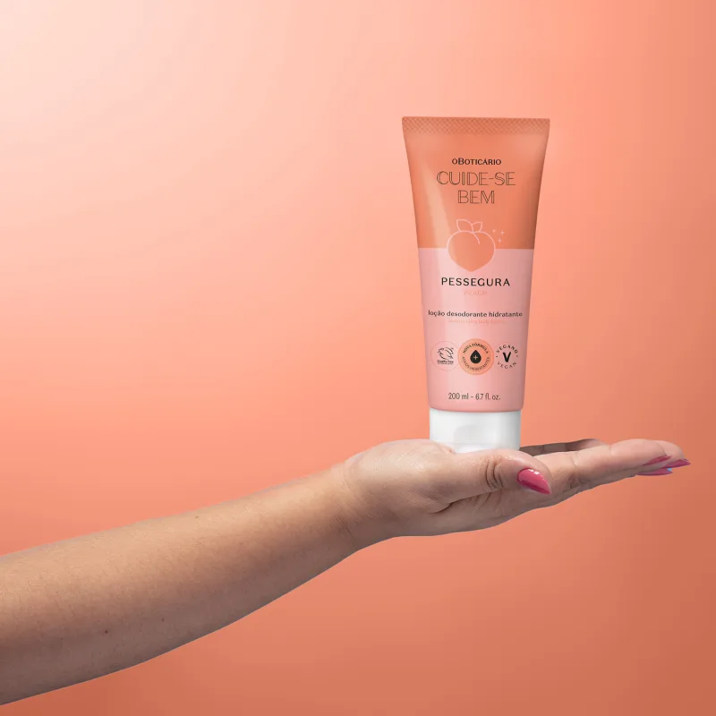Mão feminina segurando um frasco rosa e branco com pêssego ilustrado e tampa branca de loção hidratante corporal Cuide-se Bem Pessegura.