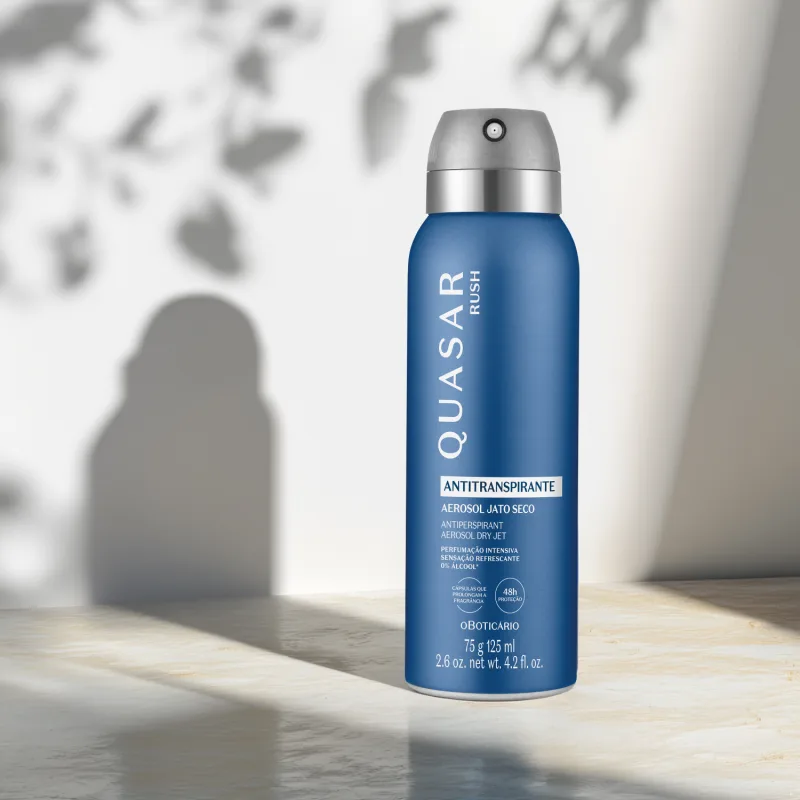 Frasco azul com rótulo e tampa nas cores prata e cinza de desodorante antitranspirante aerossol Quasar Rush. O item está em um cenário claro com sombras da embalagem e de folhagens ao fundo.