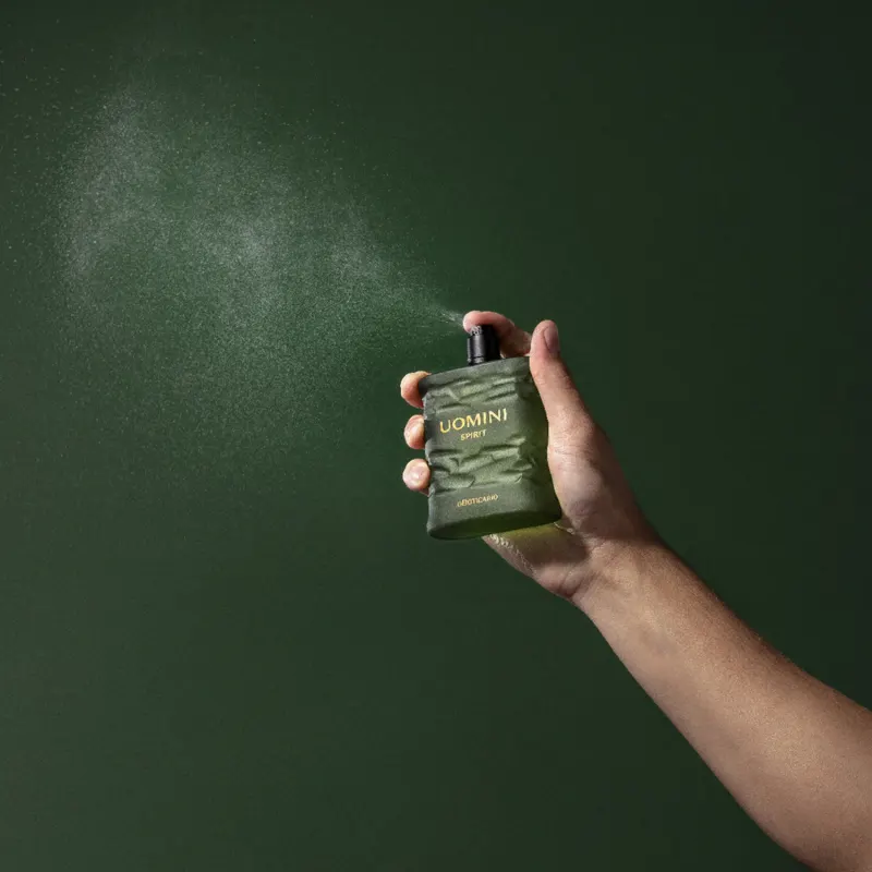 Mão segurando um frasco retangular em tom de verde oliva com tampa texturizada preta e letras laranjas contendo 100ml de desodorante colônia Uomini Spirit, espirrando o produto no ar em fundo verde bem escuro.