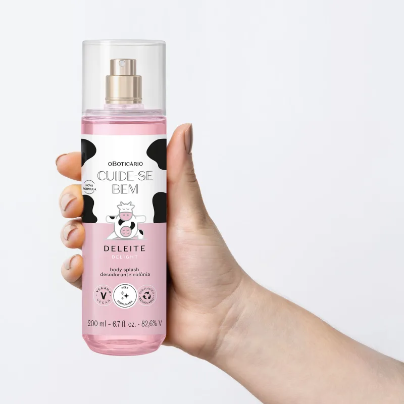 Mão feminina segurando um frasco transparente com líquido rosa, pulverizador prateado, tampa transparente e rótulo branco e rosa com manchas pretas e ilustração de vaca contendo body splash Cuide-se Bem Deleite em fundo azul.