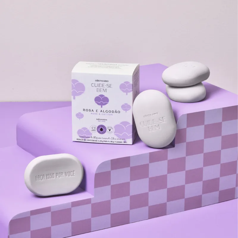 Quatro sabonetes em barra na cor lilás da marca Cuide-se Bem Rosa e Algodão ao lado de sua caixa branca e lilás em superfície em tons de roxo e fundo lilás.
