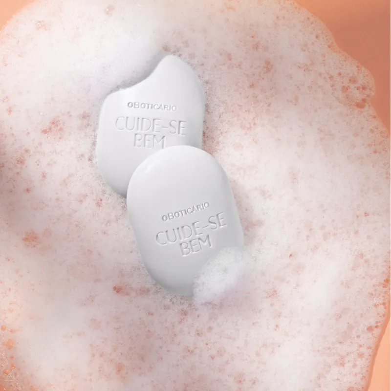 Dois sabonetes em barra ovalados brancos da marca Cuide-se Bem Nuvem em meio a muita espuma branca em fundo laranja.