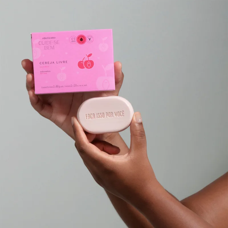 Uma mão feminina segurando uma caixa roxa com letras pretas e detalhes rosa e a outra segurando um sabonete em barra branco ovalado da marca Cuide-se Bem Cereja Livre em fundo cinza.