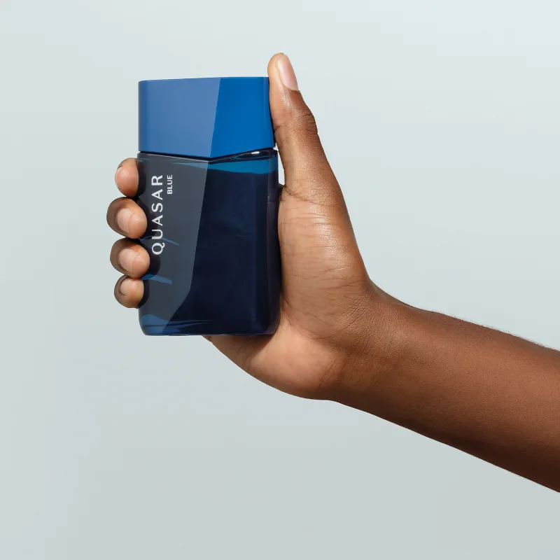 Mão masculina segurando um frasco de vidro em formato geométrico com letras amarelas contendo desodorante colônia Quasar Blue em fundo azul claro.