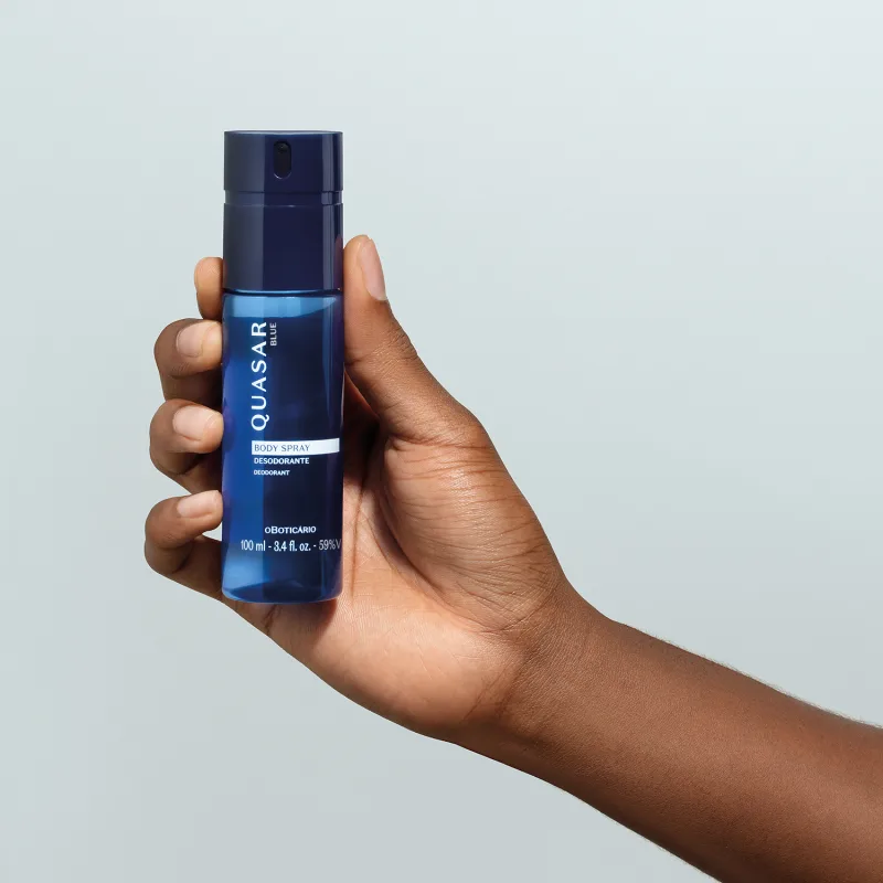 Mão masculina segurando um frasco azul marinho com letras amarelas e detalhe branco contendo body spray desodorante da marca Quasar Blue em fundo azul claro.