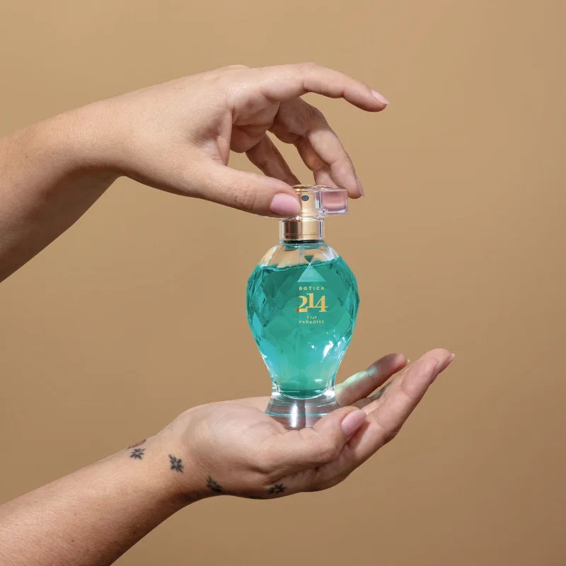 Duas mãos femininas segurando um vidro transparente lapidado com líquido azul, borrifador dourado, tampa transparente e letras douradas de Eau de Parfum Floral Especiado da marca Botica 214 Fiji Paradise em fundo marrom.