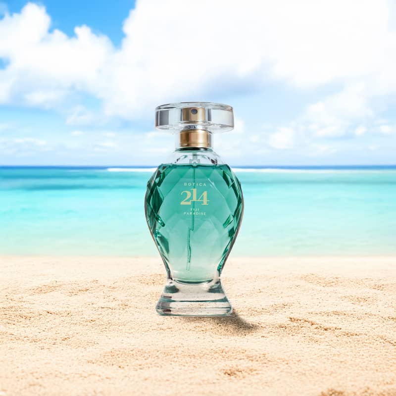 Vidro transparente lapidado com líquido azul, borrifador dourado, tampa transparente e letras douradas de Eau de Parfum Floral Especiado da marca Botica 214 Fiji Paradise em um cenário de praia, com areia, mar e céu azul.