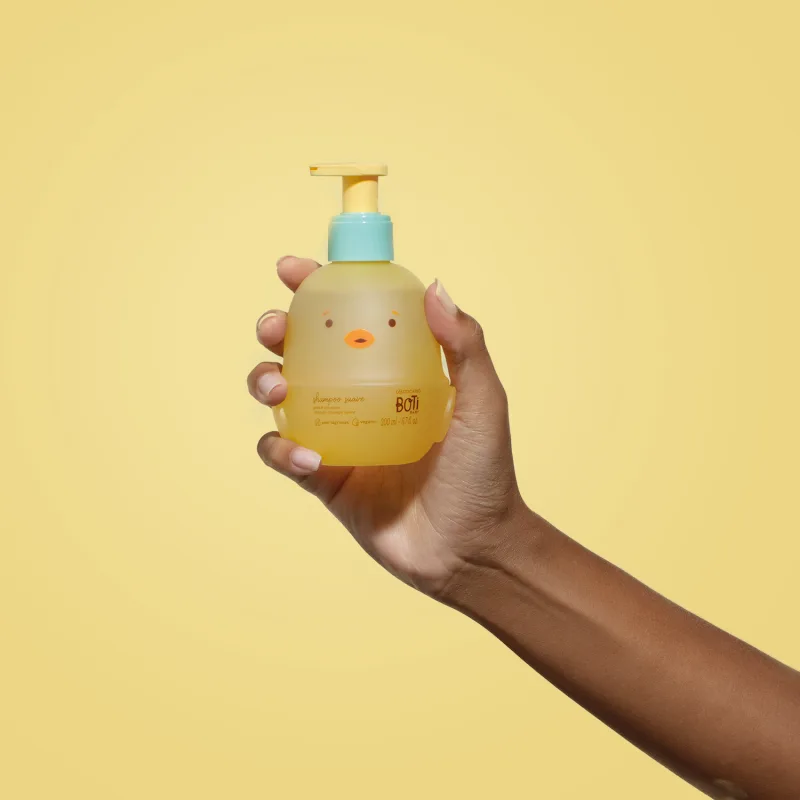 Mão segurando um frascoem forma de passarinho amarelo com detalhes laranjas, tampa azul e bomba dosadora amarela de shampoo suave da marca Boti Baby em um fundo alaranjado.