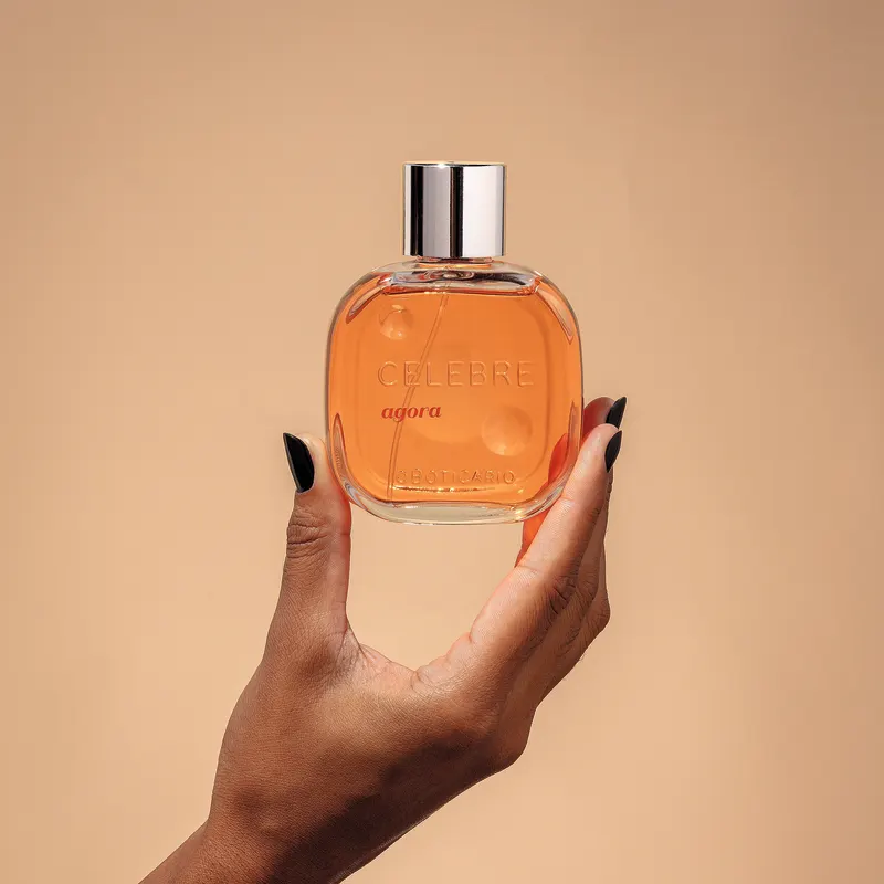 Uma mão feminina com unhas pintadas de preto segurando um frasco de vidro transparente com líquido laranja, tampa prateada e letras pretas e em relevo de desodorante colônia Celebre Agora Feminino em fundo em tons de laranja.