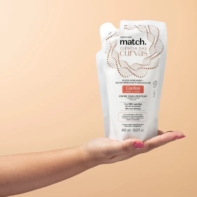 Mão segurando um Refil de creme para pentear para cabelos cacheados Match Ciência das Curvas em sachê branco com letras marrons e detalhes laranjas em fundo rosado.