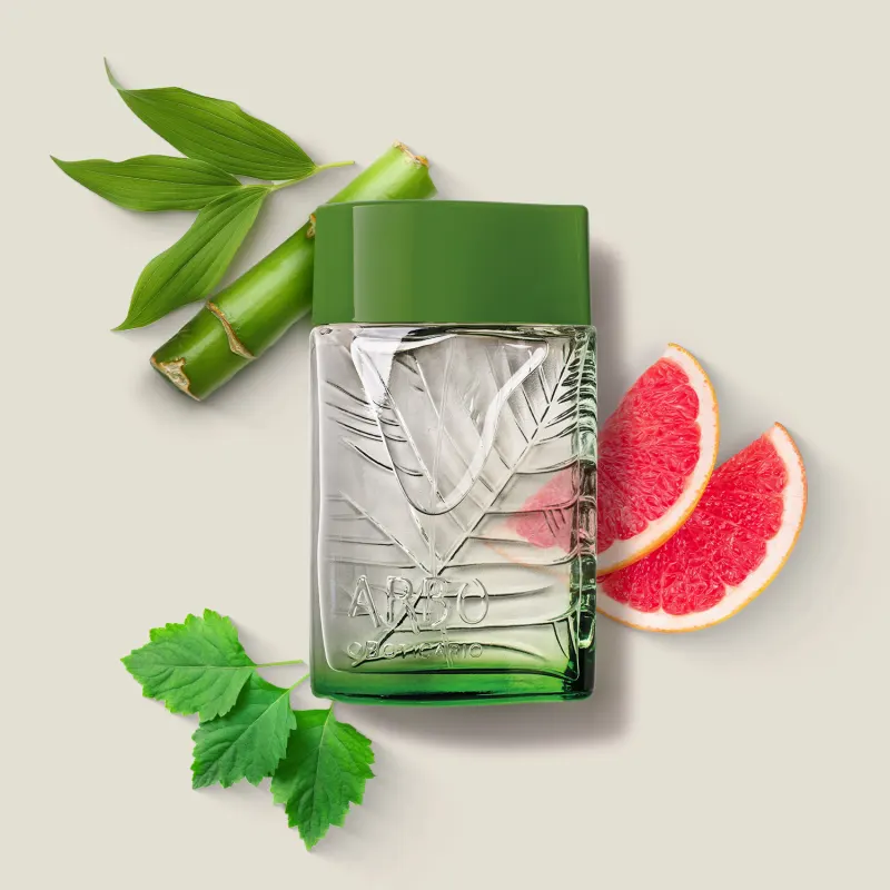 frasco verde transparente com líquido claro, folhagens em relevo e tampa verde escuro contendo desodorante colônia Arbo Puro com folhas verdes e fatias de toranja rosa ao redor, sobre superfície clara.