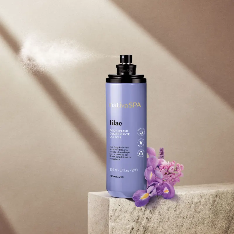Frasco cilíndrico na cor lilás com tampa preta e letras brancas e pretas  contendo body splash Nativa SPA Lilac posicionado sobre superfície de concreto, com flores lilás ao lado, em ambiente com iluminação natural.
