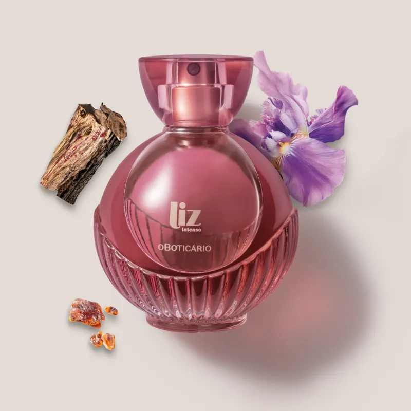 Um vidro com líquido vermelho, borrifador dourado, tampa transparente e letras douradas de desodorante colônia Liz Intenso está em um fundo cinza com flor roxa, pedaço de madeira e âmbar ao redor.