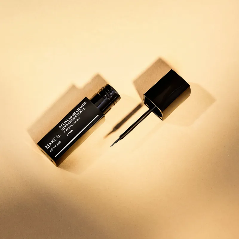 Tubo preto  com letras brancas de delineador líquido para olhos na cor preta da marca Make B., com tampa, haste e aplicador fino. A imagem tem um fundo em tons de rosa.