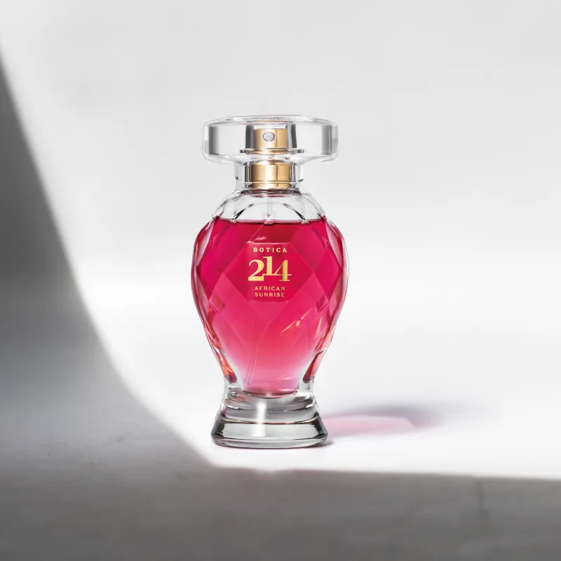 Vidro lapidado em formato de taça com líquido rosa vibrante, tampa transparente, pulverizador e letras douradas contendo 75ml de Eau de Parfum Botica 214 African Sunrise Floral Frutal em fundo claro com sombras.