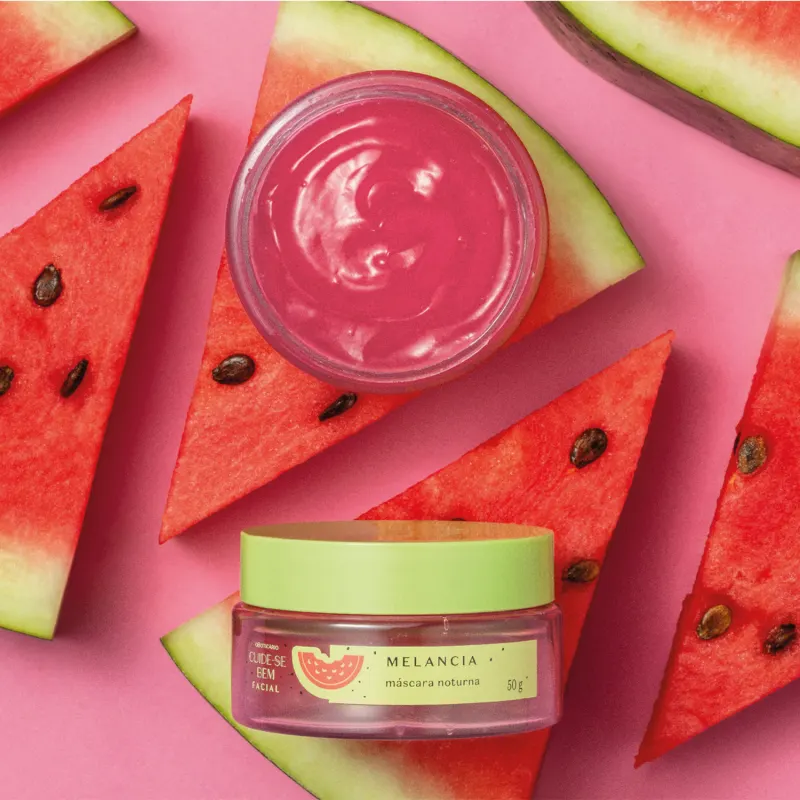 Pote com creme vermelho, tampa verde e rótulo verde com melancia ilustrada de máscara facial noturna Cuide-se Bem Feira Melancia com fatias de melancia vermelha e sementes ao redor, em um cenário de frutas e um pouco do creme em gel em cima da fruta.