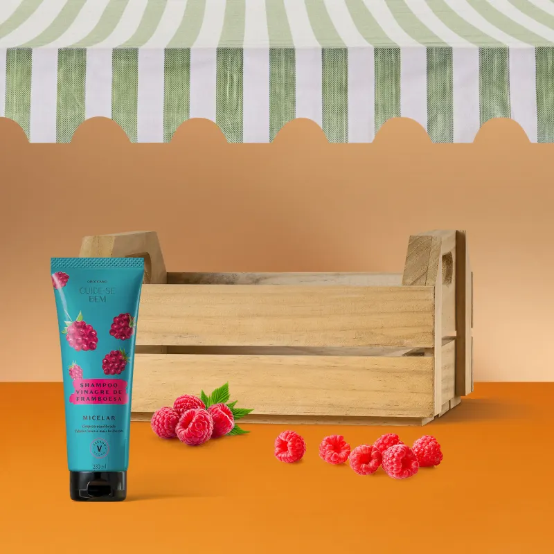 Caixa de madeira com frasco azul com tampa preta e ilustrações de framboesas na cor rosa contendo shampoo Cuide-se Bem Feira Vinagre de Framboesa rodeada por framboesas frescas sobre fundo laranja e parede listrada em verde e branco.