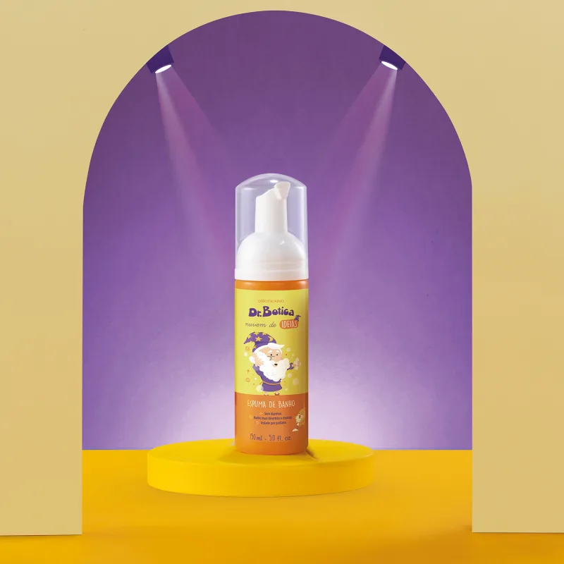 Espuma de banho corporal Nuvem de Ideias Dr. Botica em frasco com tampa branca e rótulo amarelo e laranja com ilustração do personagem da marca em destaque sobre superfície amarela, com iluminação roxa ao fundo, criando um ambiente vibrante e atraente.