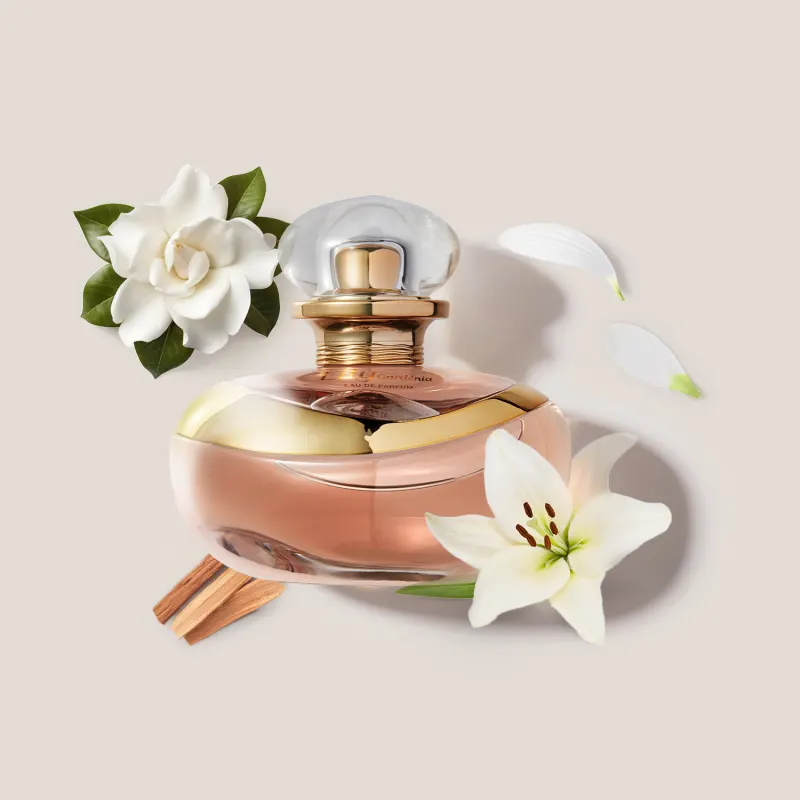 O frasco de Eau de Parfum Lily Gardênia tem formato arredondado com tampa esférica. Ao redor, há uma flor gardênia branca, pétalas de lírio branco e gravetos de canela. O conjunto transmite uma atmosfera floral
