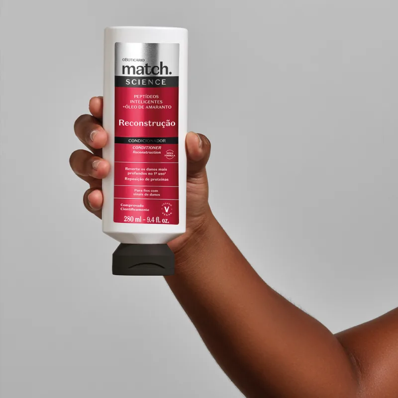 Mão segurando um vidro branco com tampa preta e rótulo vermelho, preto e prateado de condicionador Match Science Reconstrução em fundo cinza.