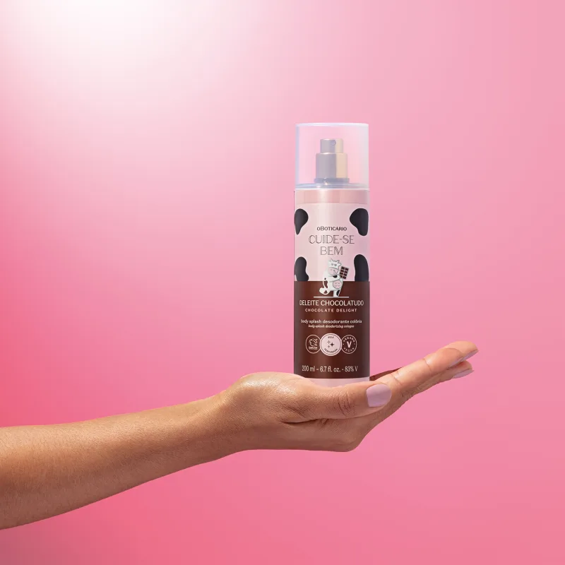 Mão feminina segurando um frasco de body splash Cuide-se Bem Deleite Chocolatudo em fundo rosa.