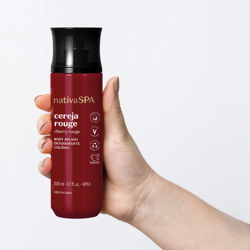 Mão feminina segurando um frasco vermelho escuro com tampa preta de body splash Nativa SPA Cereja Rouge em fundo azul claro.