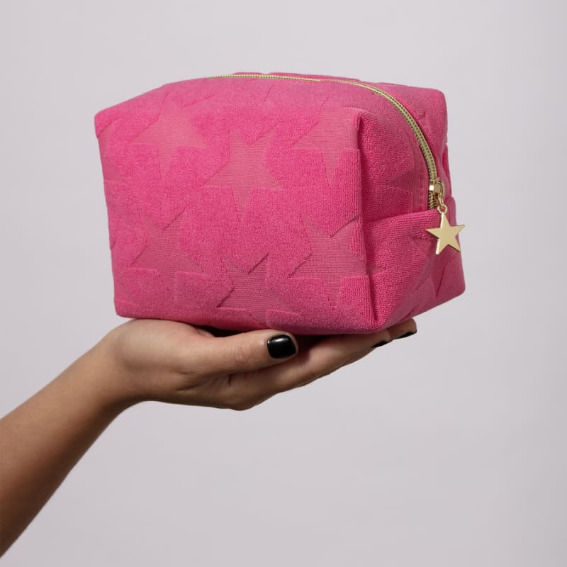 Mão segurando um nécessaire rosa macio com estampa de estrelas e fecho em zíper com pingente também de estrelas em fundo cinza claro.