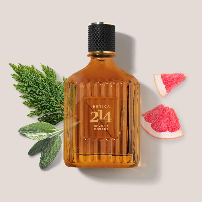 Frasco de Eau de Parfum Fougére Aromático Botica 214 Sevilla Dorada em fundo claro, com ramos de plantas verdes e fatias de toranja ao redor, criando um cenário natural e aromático.