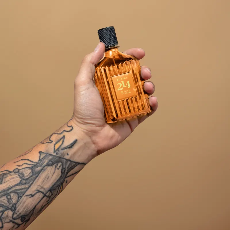 Frasco de Eau de Parfum Fougére Aromático Botica 214 Sevilla Dorada em mão com braço tatuado, sobre fundo bege, em ambiente minimalista e monocromático.
