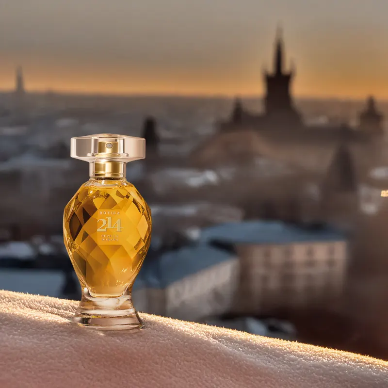 Frasco de Eau de Parfum Floral Frutal Botica 214 Sevilla Dorada em primeiro plano, com paisagem de castelo e casas ao fundo em tons de laranja e cinza durante o pôr do sol.