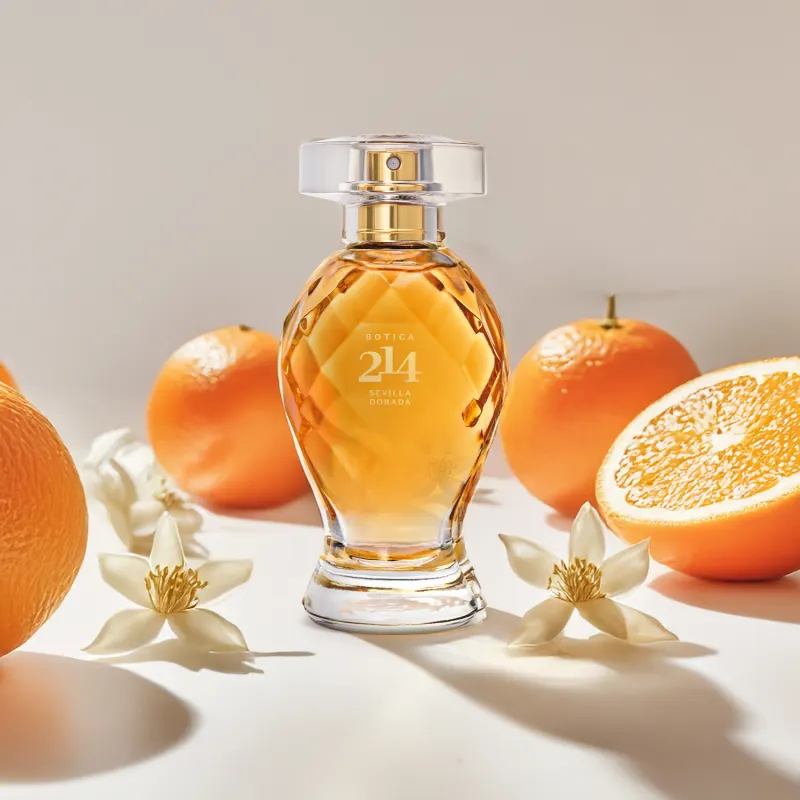 Frasco de Eau de Parfum Floral Frutal Botica 214 Sevilla Dorada em meio a laranjas frescas e flores brancas sobre superfície clara, em cenário iluminado e com cores quentes predominantes.