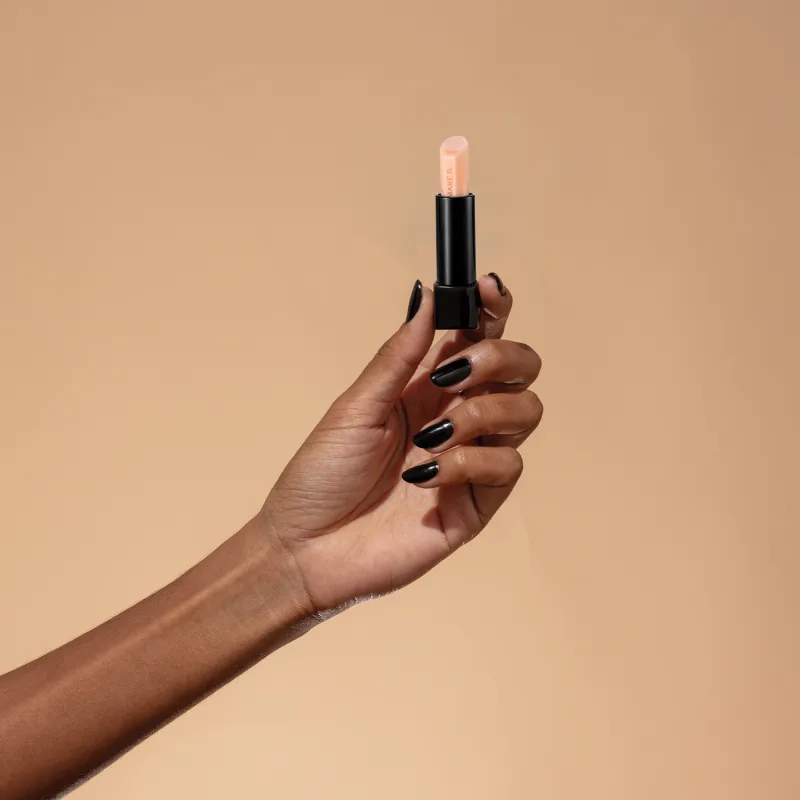Mão feminina com unhas pintadas de esmalte escuro segurando um balm labial noturno da marca Make B. Lip Chronology Night Plump sobre fundo bege.
