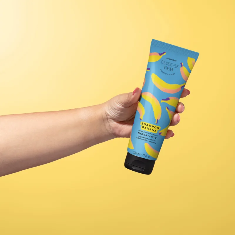Imagem de uma mão segurando um frasco de shampoo hidratante da marca Cuide-se Bem Feira Banana, em embalagem azul com bananas ilustrando, contendo 230 ml do produto. O fundo é em tons de branco e amarelo.